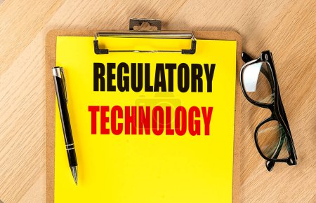 Photo for REGULATORY TECHNOLOGY text on a yellow paper on clipboard with pen and glasses. - Royalty Free Image