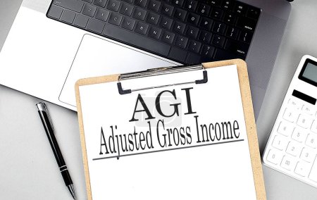 AGI -ADJUSTED GROSS INCOME Wort auf einer Zwischenablage auf dem Laptop mit Taschenrechner und Stift .