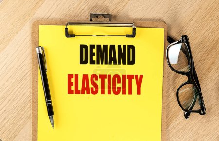 DEMAND ELASTICITY text on a yellow paper on clipboard with pen and glasses. 