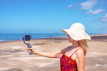 Foto de Mujer madura tomando selfies con un teléfono móvil en un trípode en la duna de la playa. Tecnología y concepto de verano - Imagen libre de derechos