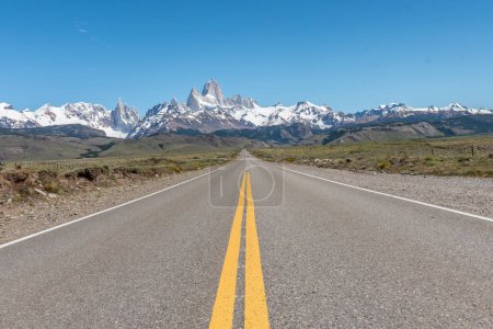 A long road with a yellow line down the middle. The road is empty, the sky is clear and the Mount Fitz Roy at the background.