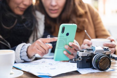 Foto de Mujer amigos mirando el teléfono celular sentado en la cafetería con cámara fotográfica, consultar mapa turístico de la ciudad - Imagen libre de derechos