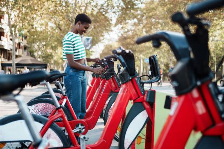 African woman taking a red bicycle in a bike rental station in the city with mobile sharing app. Ecological Urban mobility