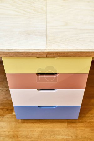 Multi-colored facades of roll-out drawers for stationery inside children table. Fragment of sawhorse desk made of plywood and solid oak upper angle view