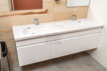 Wall mounted bathroom vanity cabinet with double white porcelain sink and silver sensor faucets in modern bathroom with concrete and wood tiles