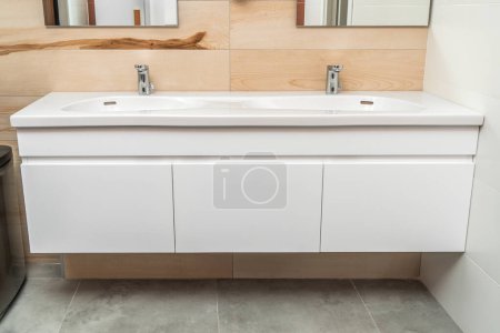 Wall mounted bathroom vanity cabinet with double white porcelain sink and silver sensor faucets in modern bathroom with concrete and wood tiles