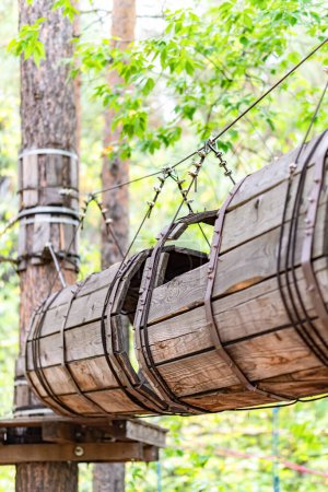 Wooden barrel tunnel obstacle suspended between trees, with cables and safety lines at adventure rope park