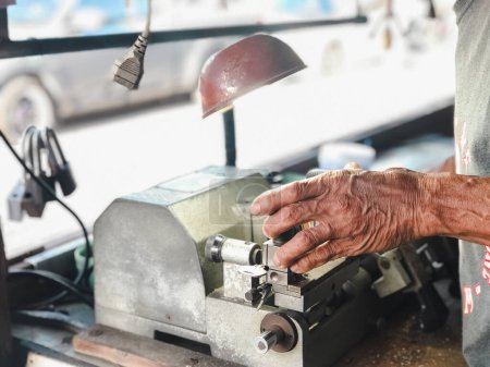 Locksmith Crafting Keys on Professional Equipment. Las manos expertas de un cerrajero trabajando en un equipo de corte de llaves bajo una luz de tarea en un taller.