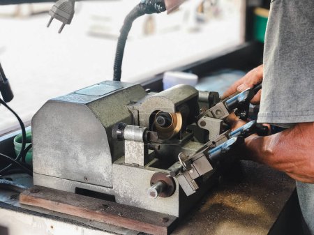 Locksmith Crafting Keys on Professional Equipment. Las manos expertas de un cerrajero trabajando en un equipo de corte de llaves bajo una luz de tarea en un taller.