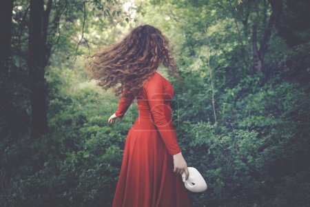 surreale Frau in Rot mit Maske in der Hand, die frei im Wald läuft, abstraktes Konzept