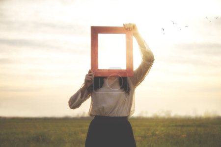 Photo for Woman holds an empty frame that makes her face disappear reflecting the sky, abstract concept - Royalty Free Image