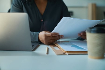 Femme multiraciale assis à atable sur le lieu de travail en face d'un ordinateur portable et tient des documents de travail dans ses mains