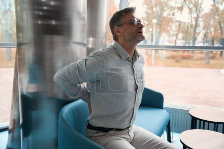 Photo for Gray-haired man in glasses is located on a sofa in a recreation area, his back hurts - Royalty Free Image