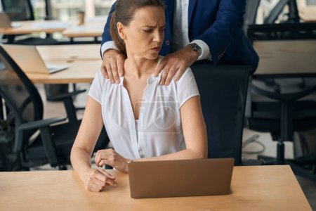 La mujer está usando la computadora portátil en el lugar de trabajo mientras el hombre está haciendo su masaje y coqueteando mientras ella se siente abusada