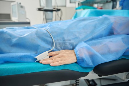 Foto de Paciente en traje protector azul acostado con oxímetro de pulso en el dedo en el sofá médico en el quirófano - Imagen libre de derechos