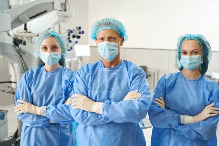 Foto de Grupo de médicos profesionales en traje de protección, máscara y sombrero de pie en la sala de operaciones en la clínica - Imagen libre de derechos