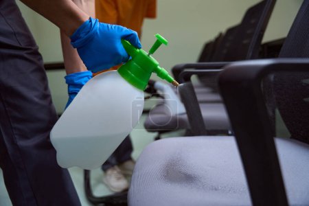 Foto de Foto recortada de limpiador en guantes sosteniendo la botella de desinfectante y rociándola sobre los muebles - Imagen libre de derechos