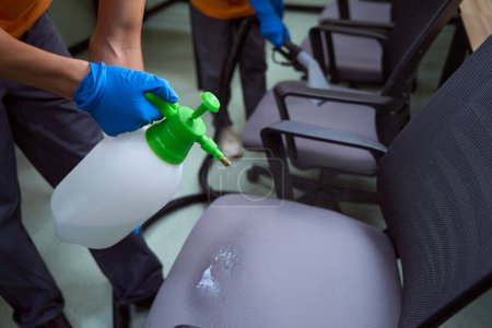 Téléchargez les photos : Plan recadré de deux employés de l'entreprise de nettoyage pulvérisant du liquide désinfectant sur des chaises - en image libre de droit