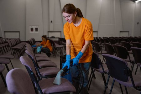 Photo for Pleasant young woman in rubber gloves carefully vacuum-cleaning chairs in sitting area while her collegue tidying up in the background - Royalty Free Image