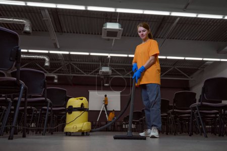 Photo for Full-size photo of focused young janitor vacuum-cleaning the floor in the conference hall - Royalty Free Image
