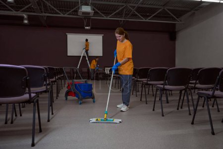 Foto de Conserje profesional lavando el piso con una fregona mientras sus colegas limpian muebles en el fondo - Imagen libre de derechos