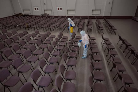 Photo for Two professional cleaners in overall protective garments spraying chemicals for disinfection on chairs in conference room - Royalty Free Image