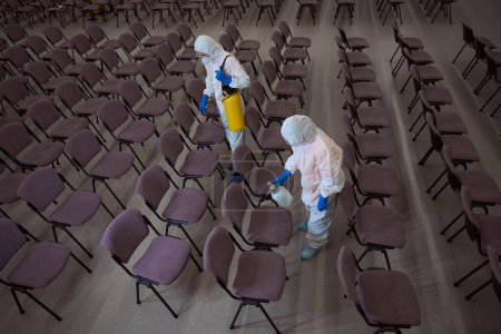 Photo for Dedicated cleaning service workers spraying disinfectant on chairs in the conference hall - Royalty Free Image