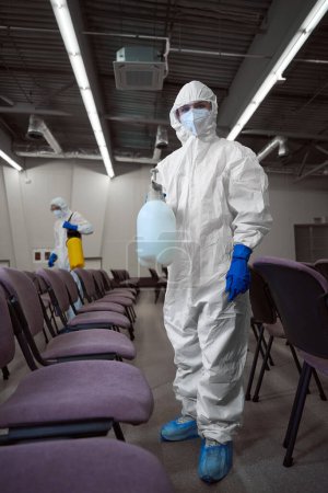 Photo for Full-size photo of well-trained cleaner in overall protective gear posing with sanitation chemicals in the middle of hall sitting area - Royalty Free Image