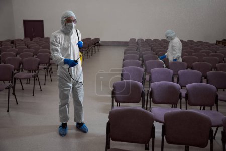 Téléchargez les photos : Nettoyants certifiés dans les vêtements de protection et masques pulvérisation de désinfectant liquide sur les chaises dans le bâtiment - en image libre de droit