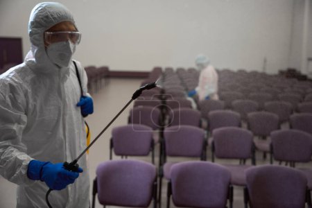 Photo for Waist-up photo of person in protective overalls and mask spraying disinfecting liquid while collegue sanitizing in the background - Royalty Free Image