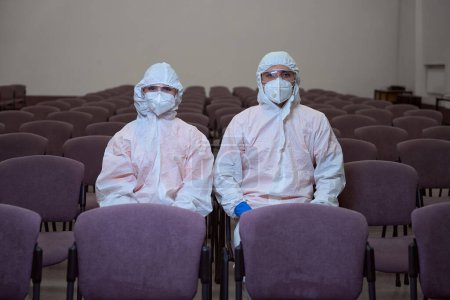 Photo for Two people in full-body protective suits, masks and goggles sitting together on chairs and looking at the camera - Royalty Free Image