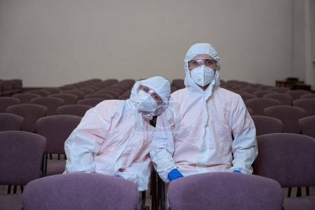 Photo for Two hard-working janitors in full-body protective gear resting while sitting on chairs next to each other - Royalty Free Image