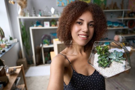 Photo for Adorable young lady smiling at the camera while holding a beautiful glass terrarium with small plants - Royalty Free Image