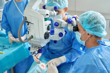 Photo for Team of doctors sitting near patient lying on couch and doing an eye operation, looking in surgical microscope - Royalty Free Image