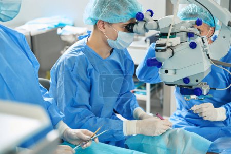 Foto de Joven médico en traje protector, máscara y sombrero de pie cerca del dispositivo con prismáticos y mirando a través de él en el paciente - Imagen libre de derechos