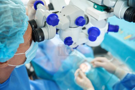 Foto de Trabajador médico en protección, máscara y sombrero mirando en el microscopio quirúrgico y mirando a través de él en el paciente - Imagen libre de derechos