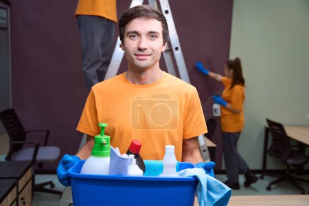 Photo for Waist-up photo of young man holding a box with cleaning supplies and looking at the camera - Royalty Free Image