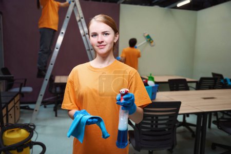 Photo for Beautiful young woman in rubber gloves holding spray bottle and wipe while cleaning team is working on the apartment - Royalty Free Image