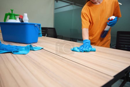 Foto de Foto recortada de limpiador certificado en guantes usando tela y rociando productos químicos mientras se desinfecta la mesa - Imagen libre de derechos