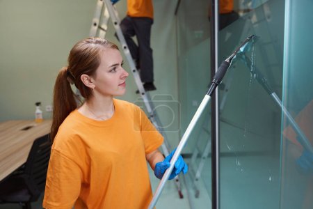 Téléchargez les photos : Jeune femme concentrée utilisant des outils de nettoyage de verre intérieur tout en essuyant la fenêtre - en image libre de droit