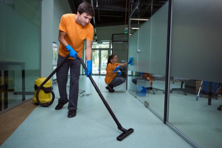 Téléchargez les photos : Jeune homme aspirant le sol tandis que sa collegue nettoyer la surface de verre dans le bâtiment - en image libre de droit