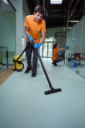 Enthusiastic janitor using the vacuum-cleaner on the floor while his partner wiping the glass surface