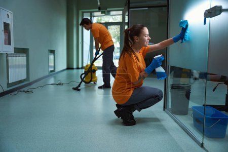 Téléchargez les photos : Joyeux femme concierge nettoyage de la surface de verre tandis que son partenaire aspirateur le sol - en image libre de droit