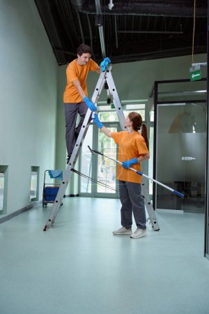 Photo for Young woman passing cleaning supplies to man on a step-ladder while helping clean the building - Royalty Free Image