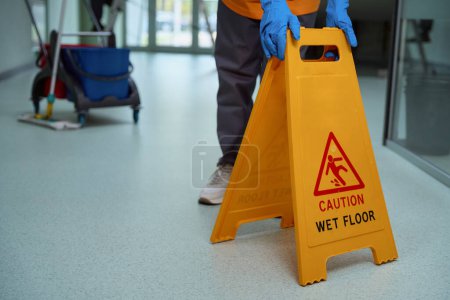 Photo recadrée de nettoyant dans des gants plaçant un panneau d'avertissement jaune sur le sol fraîchement nettoyé