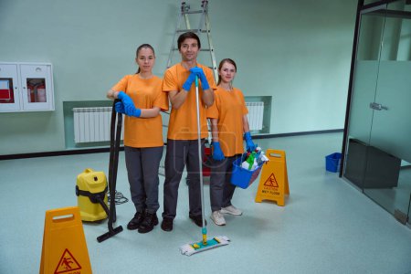 Téléchargez les photos : Jeunes employés joyeux du service de nettoyage posant avec différentes fournitures ménagères et regardant la caméra - en image libre de droit