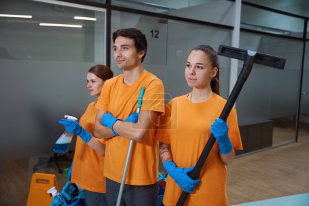 Foto de Tres limpiadores profesionales en camisas y guantes a juego que sostienen fregonas y productos químicos - Imagen libre de derechos