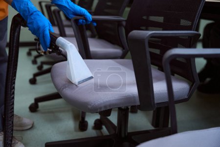 Téléchargez les photos : Photo recadrée du concierge dans des gants en caoutchouc nettoyant le rembourrage sur des chaises avec aspirateur - en image libre de droit