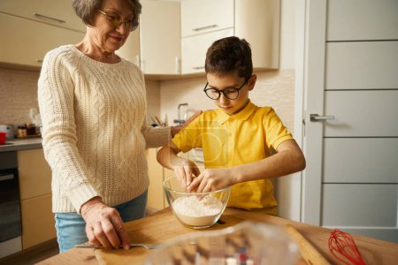 Foto de Niño de pie cerca de la mesa en la cocina y sosteniendo huevo de gallina, abuela de pie cerca y mirando al niño - Imagen libre de derechos