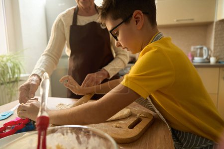 Foto de Niño de pie cerca de la mesa en la cocina, rollos de masa con rodillo, abuela ayuda cerca - Imagen libre de derechos